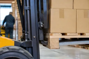 Wózek widłowy transportuje paletę z kartonami w magazynie, w tle rozmyta postać pracownika.